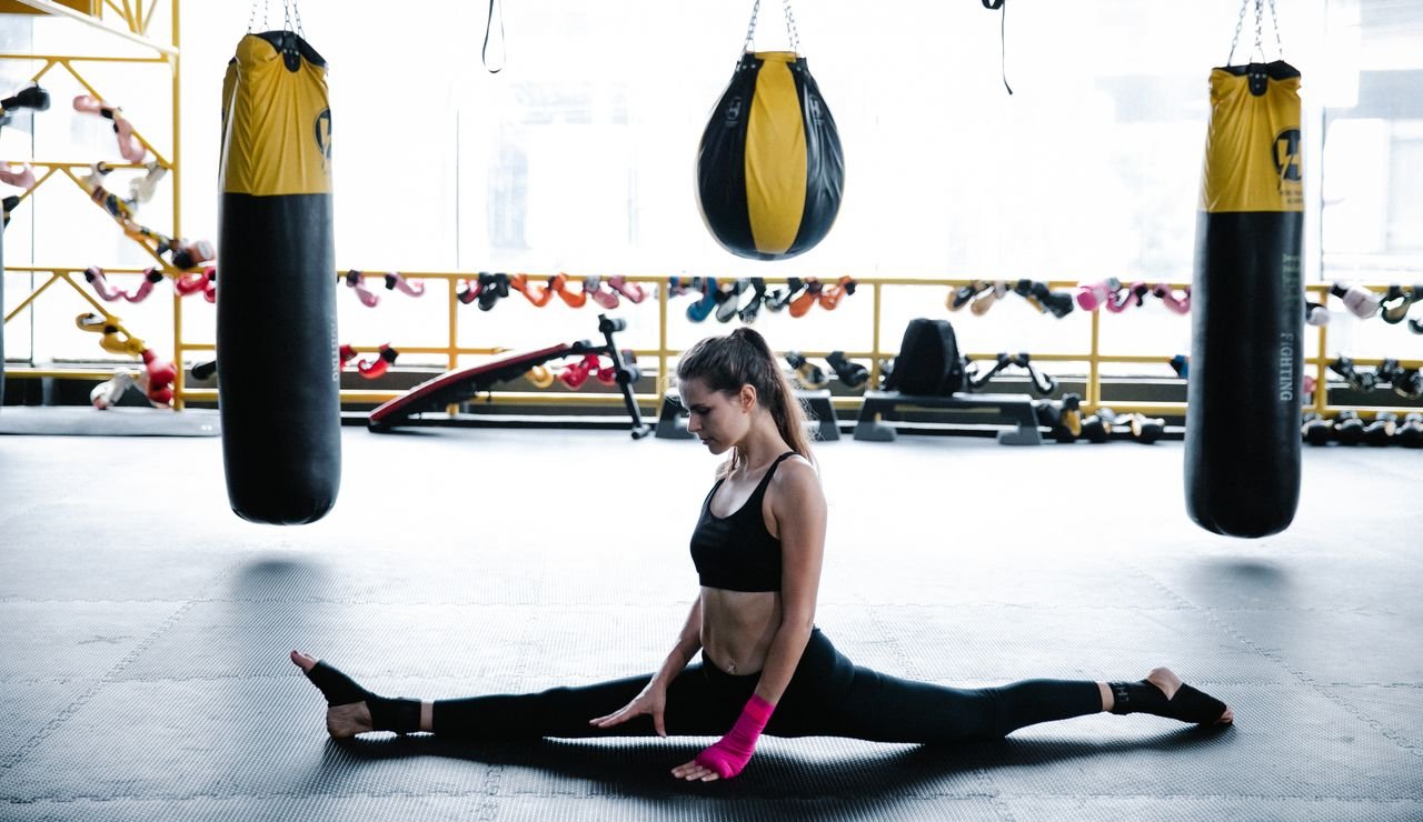 A young woman in sports attire performs splits in a gym with punching bags, showcasing flexibility and fitness.